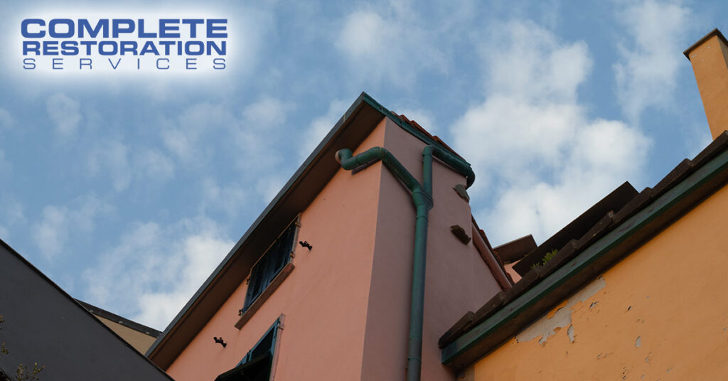 Corner of a peach-colored building with green pipes against a cloudy blue sky.