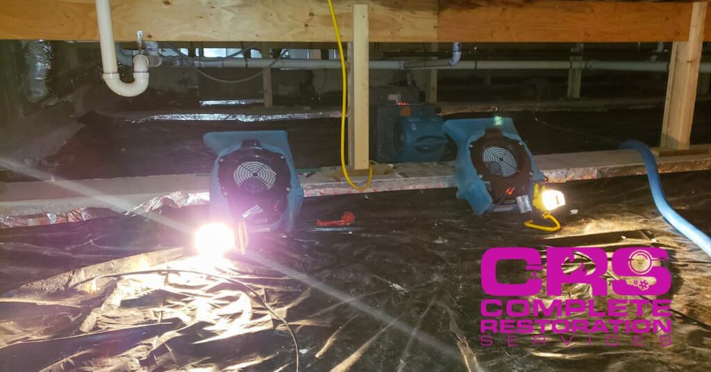 Two blue air movers with lights on in a crawl space with black plastic covering the ground.