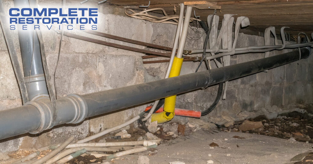 Pipes and cables running along a concrete crawl space wall with debris on the ground.