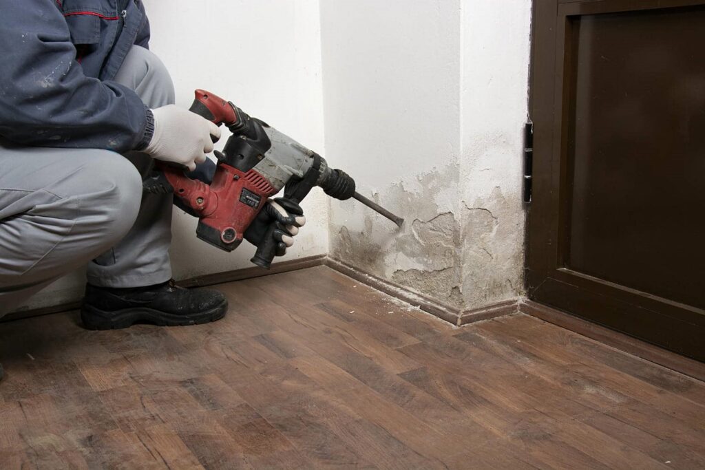 Person using a power drill to remove damaged plaster from a wall corner near a door.