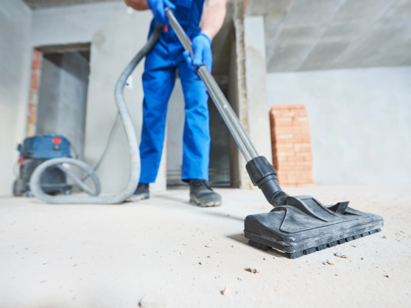 Person in blue coveralls vacuuming dust from a concrete floor in an unfinished room. construction cleanup services