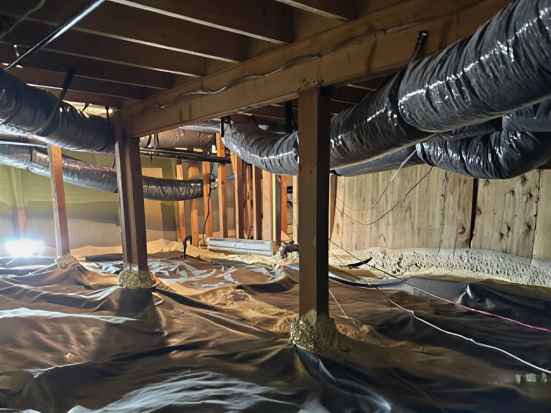 Crawl space with wooden beams, insulated ductwork, and a plastic vapor barrier on the ground. crawl space remediation