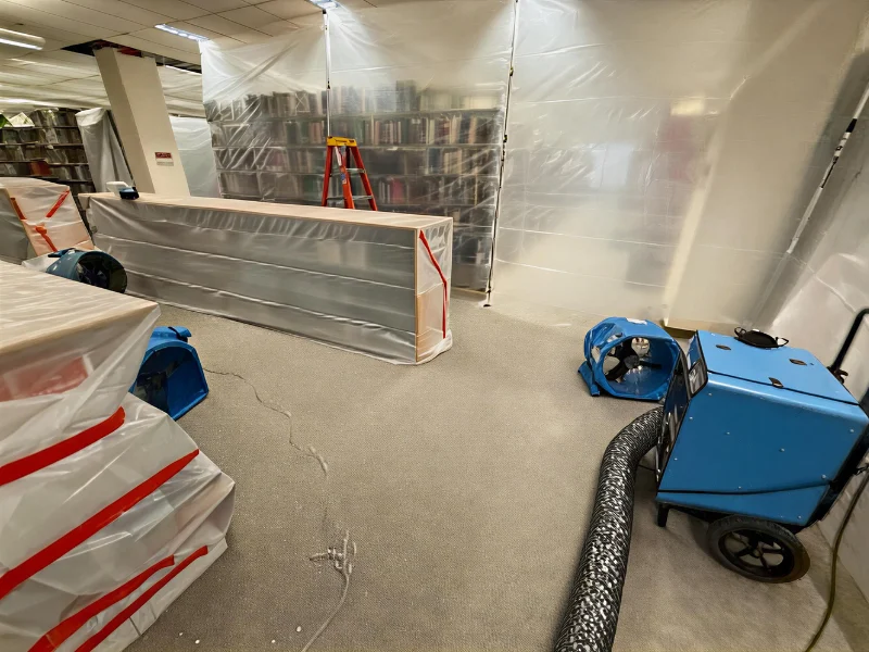 Library shelves and furniture covered in plastic with air scrubbers and a ladder in a renovation area. commercial restoration services