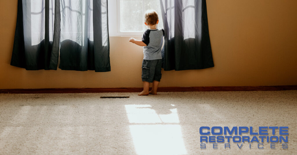 A toddler stands barefoot on a carpeted floor looking out a window with black curtains.