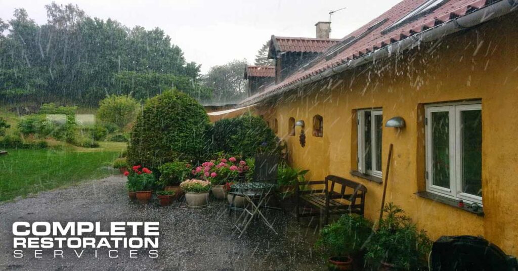 Yellow house with garden plants and outdoor furniture during heavy rain.