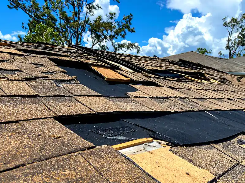 Roof with damaged and missing shingles under a blue sky with scattered clouds. storm damage restoration
