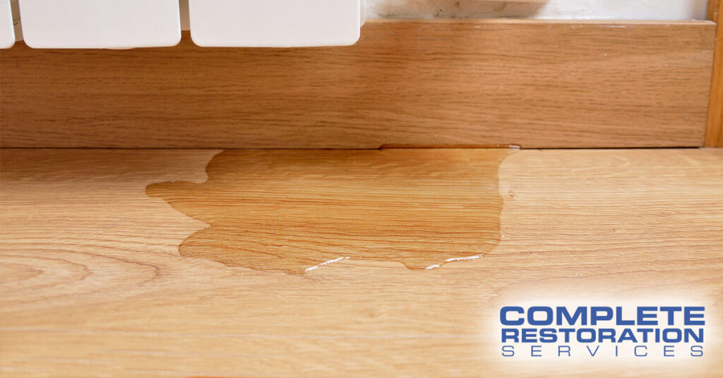 Water spill on wooden floor near baseboard heater with "Complete Restoration Services" text in corner.