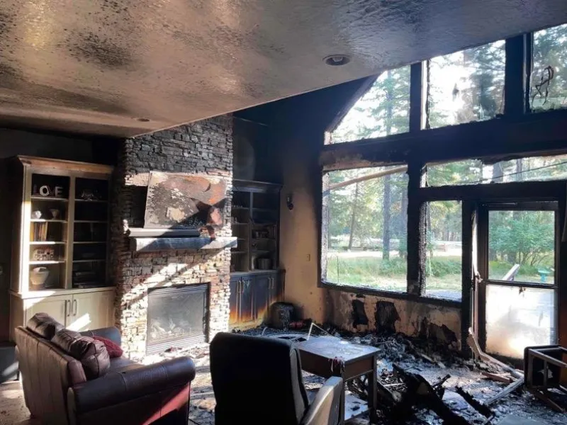 Living room with fire damage, showing charred walls, ceiling, and furniture near a stone fireplace and large windows. fire damage restoration