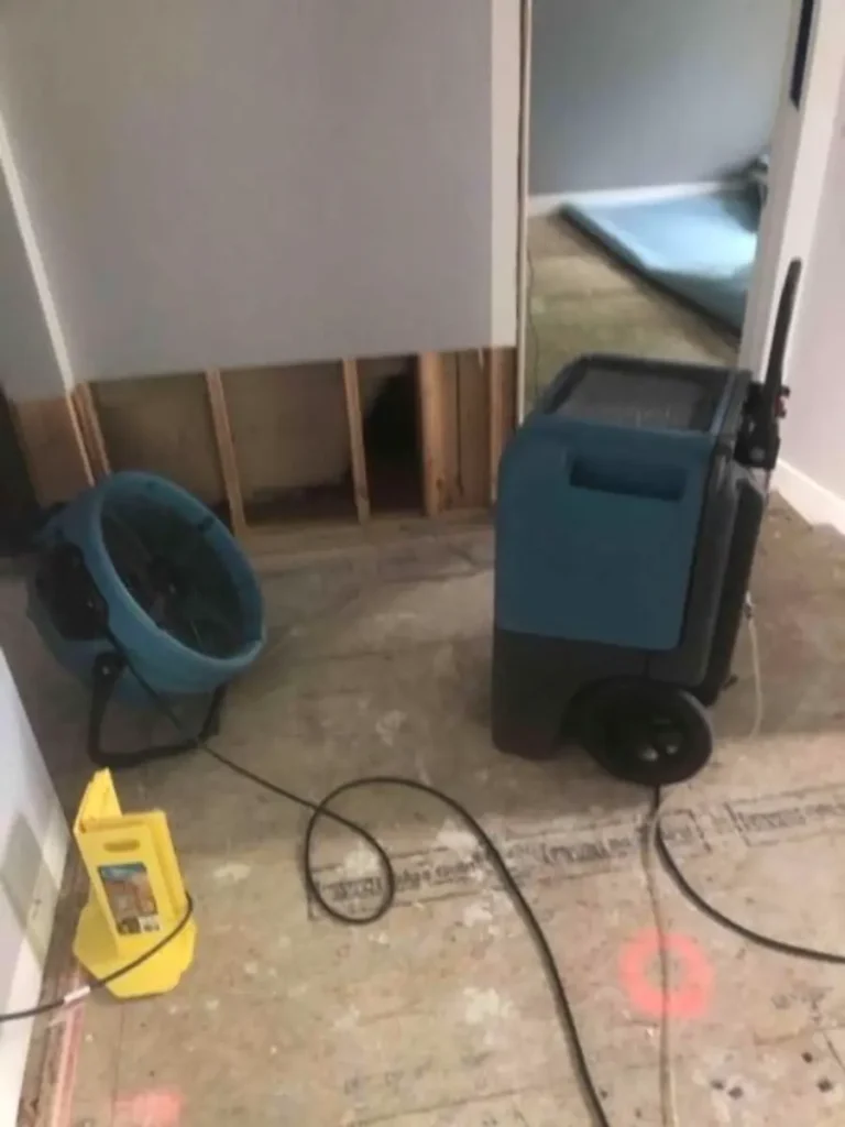 Water damage drying equipment with a fan and dehumidifier in a room with exposed lower wall panels. water damage restoration