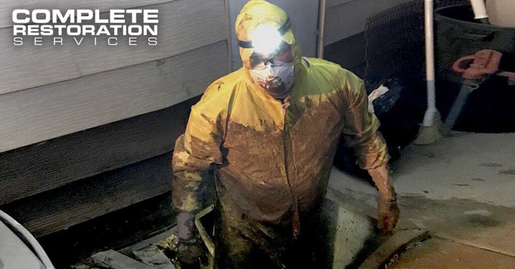 Worker in a yellow protective suit and headlamp emerging from a dirty crawl space.